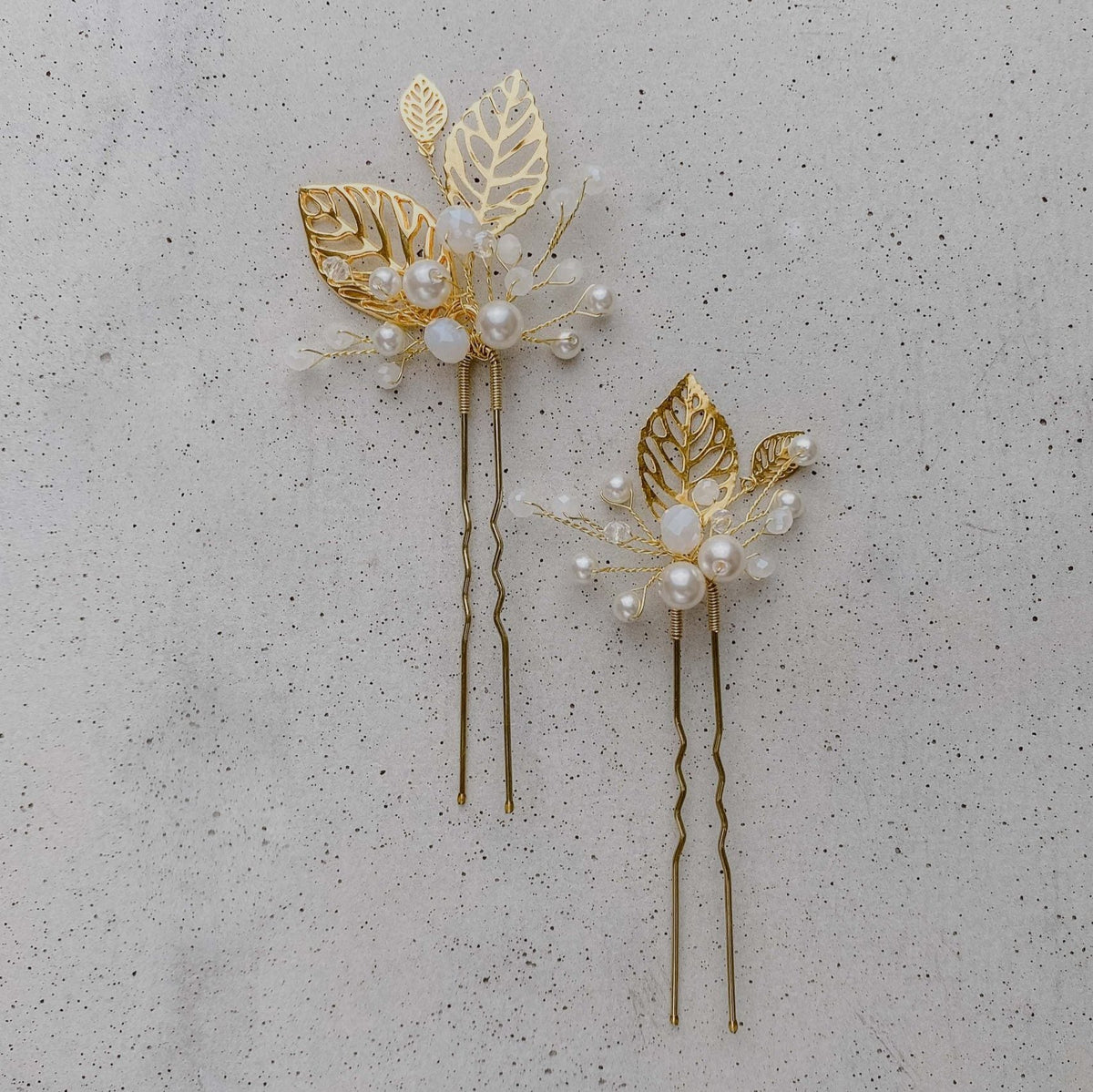 2er Set Braut Haarnadeln Zoe in Gold mit filigranen Blättern, veganen Perlen und white opal Kristallperlen, liegend auf einer Betonplatte