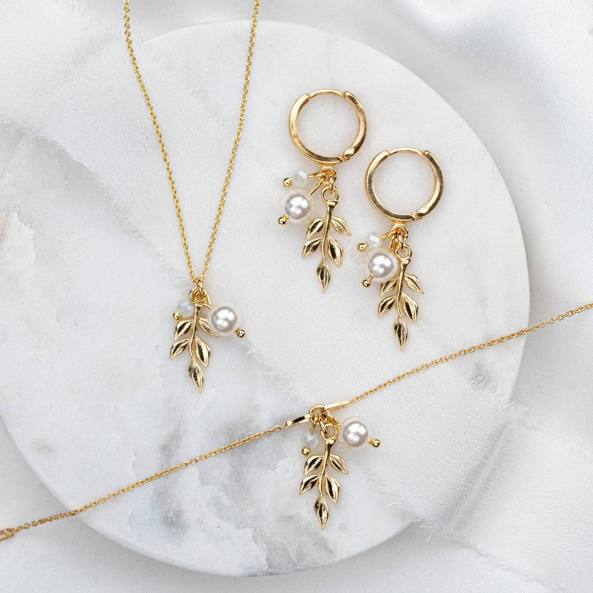 3-teiliges Schmuckset in 925er Silber 18K vergoldet bestehend aus Halskette, Creolen und Armband mit filigranen Blattanhängern, kleinen weißen Perlen und white opal Kristallperlen arrangiert auf hellem Marmorhintergrund