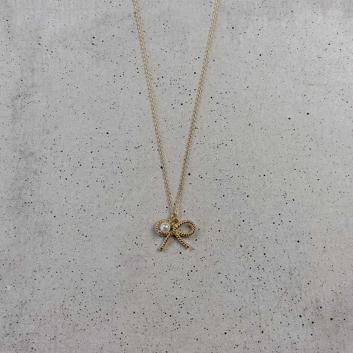 Braut Halskette Pinella aus 925 Sterlingsilber 18K vergoldet mit Schleife und veganer Perle, liegend auf Betonplatte