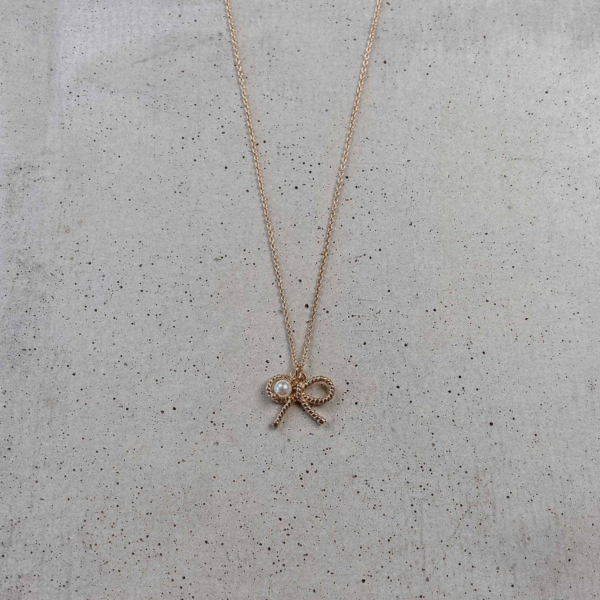 Braut Halskette Pinella aus 925 Sterlingsilber 18K rosévergoldet mit Schleife und veganer Perle, liegend auf Betonplatte