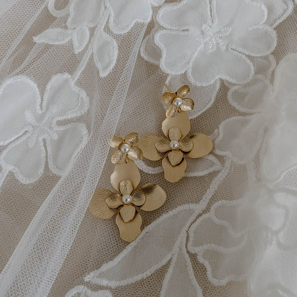 Statement Ohrringe Vika, eine große Blüte hängt an einem kleinen Blütenstecker mit veganen Perlen in 925 Sterlingsilber 18 K vergoldet, liegend auf Spitzenstoff