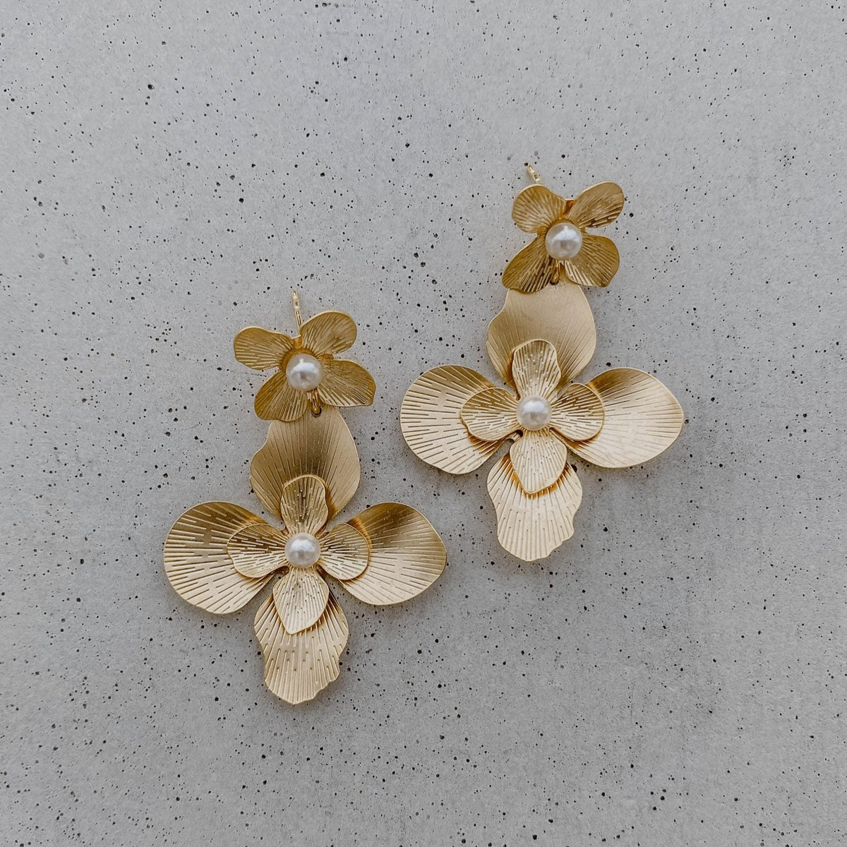 Statement Ohrringe Vika, eine große Blüte hängt an einem kleinen Blütenstecker mit veganen Perlen in 925 Sterlingsilber 18 K vergoldet, liegend auf einer Marmorplatte