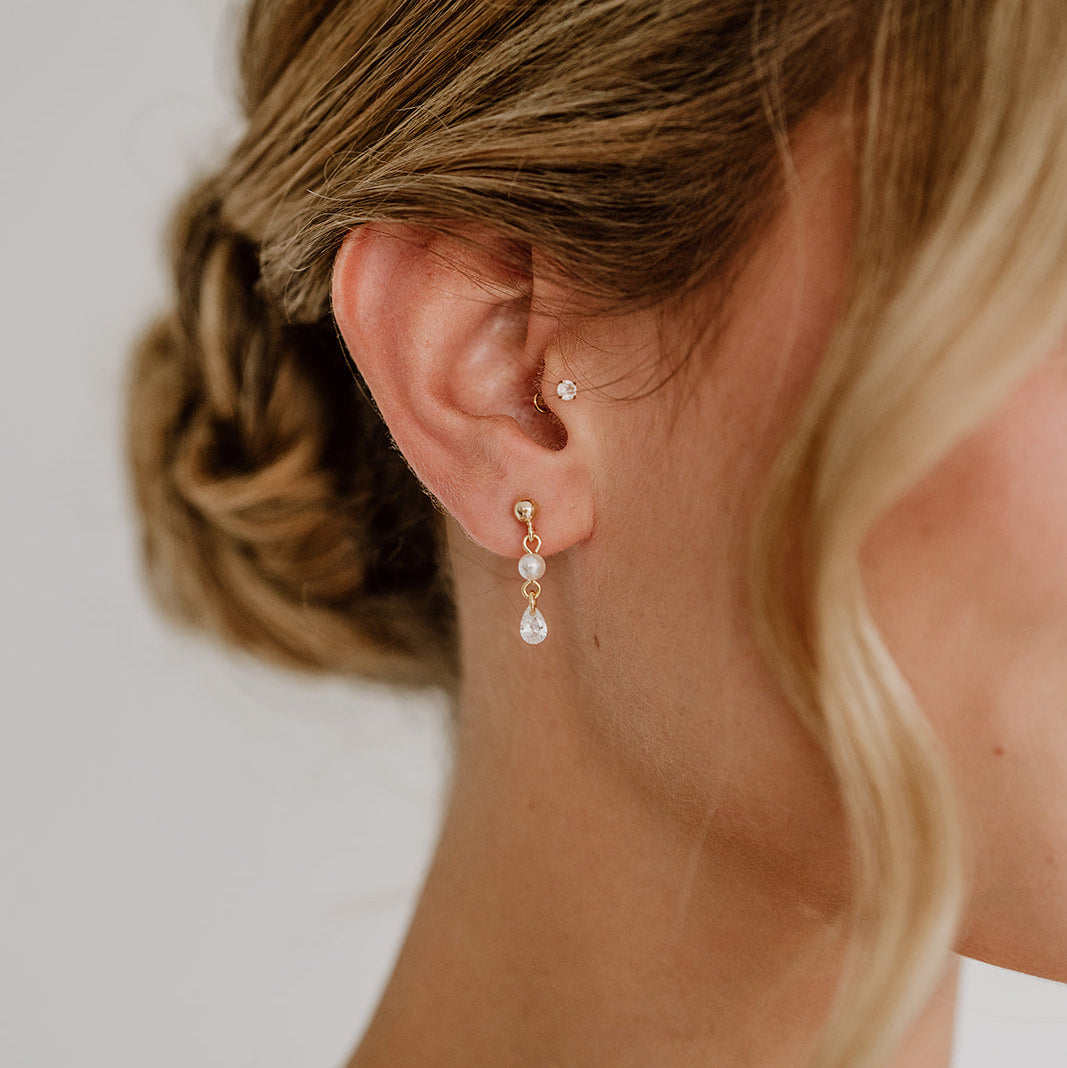 Zarte Brautohrringe aus 925er Sterlingsilber 18K vergoldet mit hängendem tropfenförmigem Zirkonia Kristall und kleiner runder Perle, elegant getragen von einem Model am Ohr und harmonisch kombiniert mit einer Hochsteckbrautfrisur.