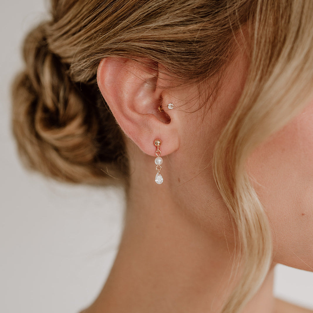 Zarte Brautohrringe aus 925er Sterlingsilber 18K rosévergoldet mit hängendem tropfenförmigem Zirkonia Kristall und kleiner runder Perle, elegant getragen von einem Model am Ohr und harmonisch kombiniert mit einer Hochsteckbrautfrisur.