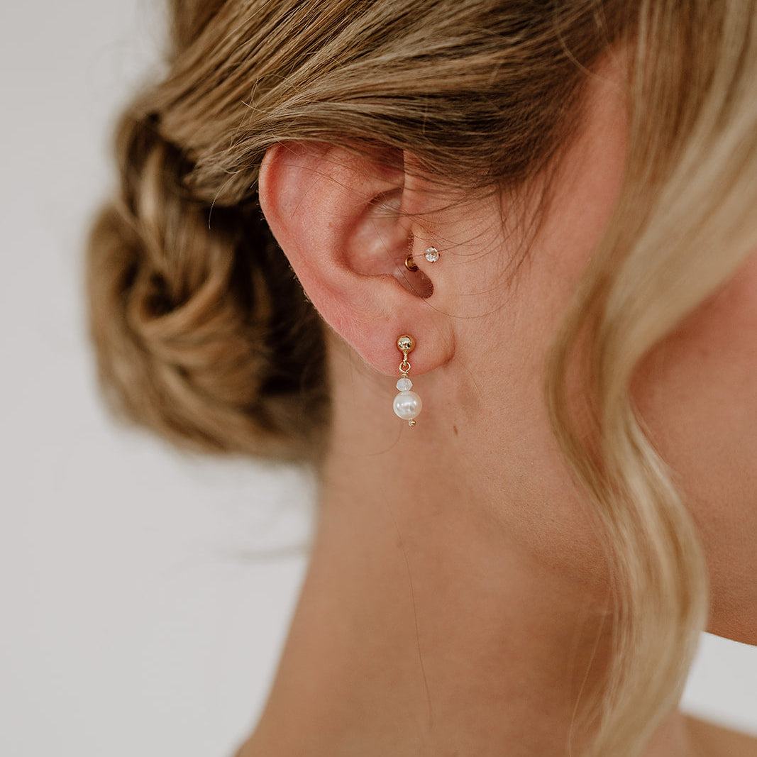 Feine Brautohrringe aus 925er Sterlingsilber 18K vergoldet. Kugelohrstecker mit kleiner Kristallperle und darunter hängender runder Perle, elegant getragen von einem Model am Ohr und harmonisch kombiniert mit einer klassischen Brautfrisur.