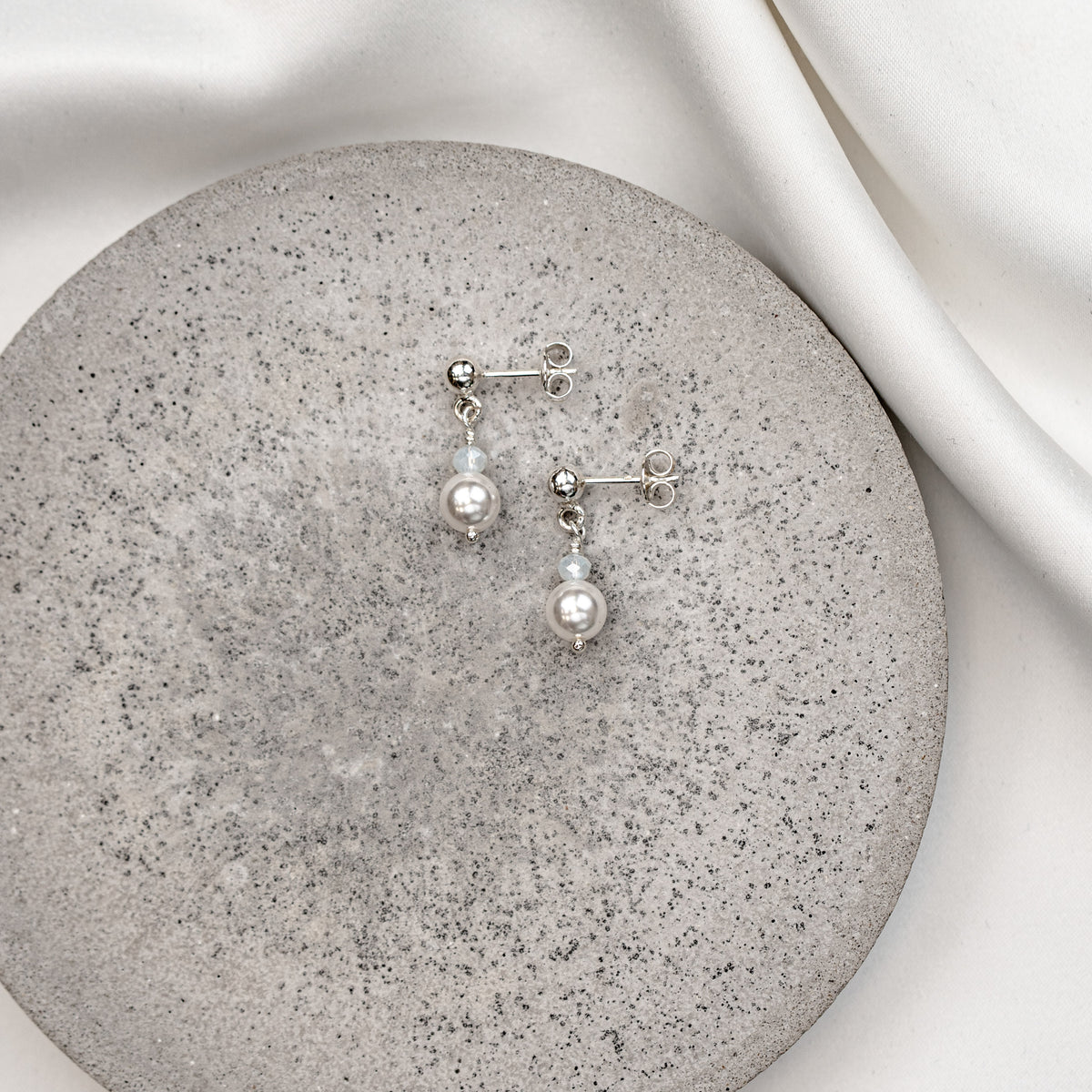 Feine Brautohrringe aus 925er Sterlingsilber. Kugelohrstecker mit kleiner Kristallperle und darunter hängender runder Perle, stilvoll arrangiert auf einer grauen Betonoberfläche.