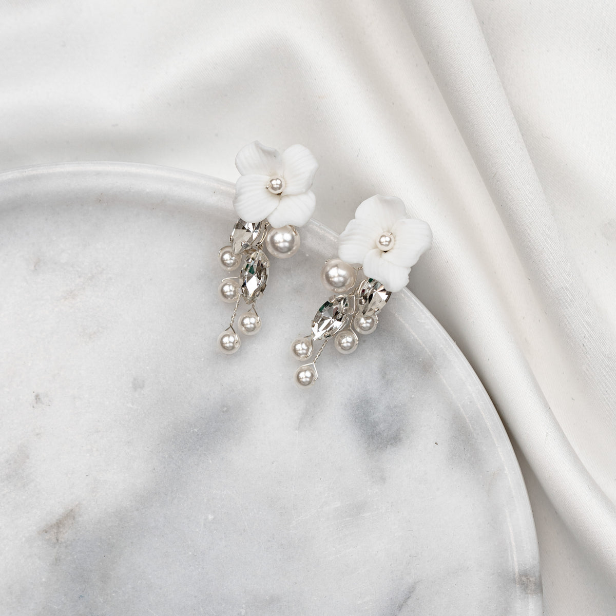 Elegante Brautohrringe aus 925er Sterlingsilber mit runder veganer Perle und darunter hängendem, blattförmigem Zirkonia Kristall, stilvoll arrangiert auf einer runden, hellen Marmorplatte.