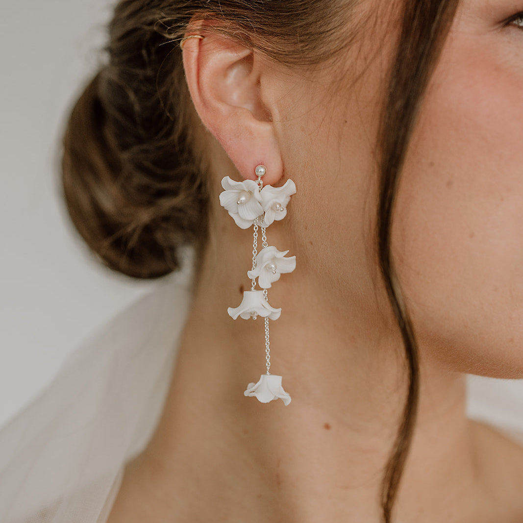 Elegante Brautohrringe aus 925er Sterlingsilber mit filigranen Porzellanblüten und zarten, beweglich hängenden Blütenelementen an feinen Kettchen, stilvoll getragen von einem Model am Ohr und harmonisch kombiniert mit einem klassischen Brautlook.