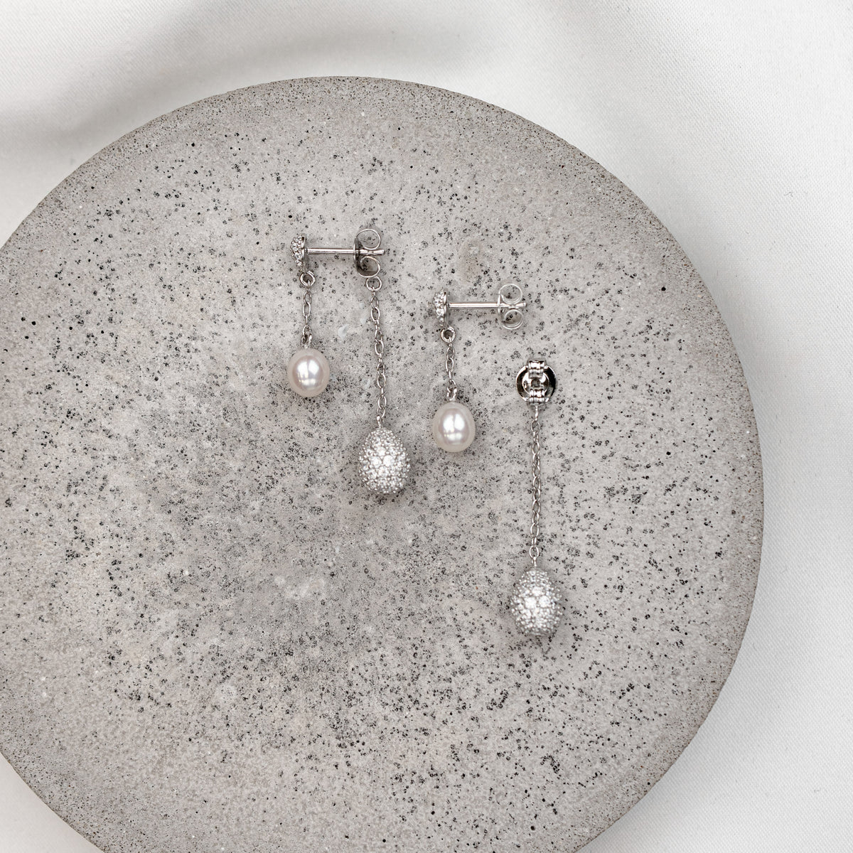Brautohrringe Elaine aus 925 Sterlingsilber mit echter Süßwasserperle und einem Tropfen besetzt mit Zirkonia Kristallen an einem feinen Kettchen, handgefertigt, liegend auf einer Betonplatte