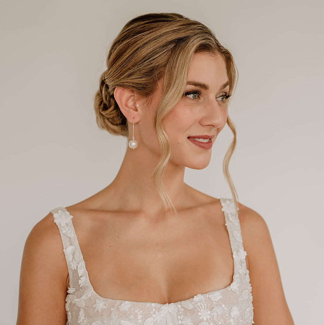 Feine Brautohrringe aus 925er Sterlingsilber mit runder veganer Perle an einem schlichten, filigranen Haken, elegant getragen von einem Model am Ohr und harmonisch kombiniert mit einem weißen, floral verzierten Brautkleid und einer klassischen Brautfrisur.