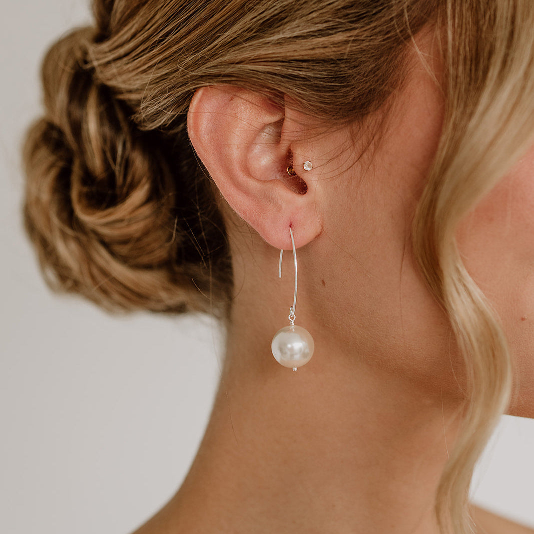 Feine Brautohrringe aus 925er Sterlingsilber mit runder veganer Perle an einem schlichten, gebogenen Haken, elegant getragen von einem Model am Ohr und harmonisch kombiniert mit einer klassischen Brautfrisur.