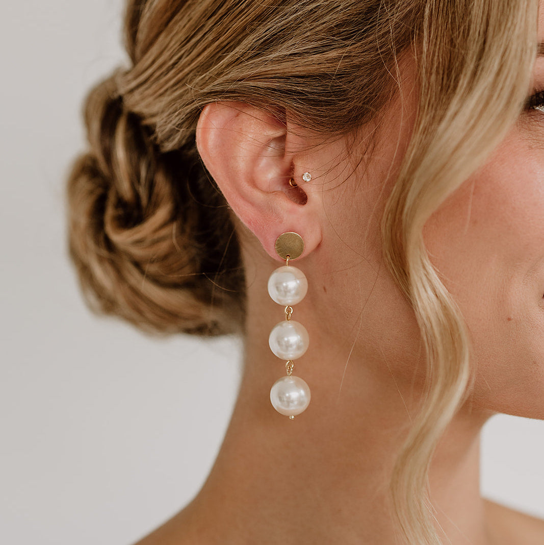 Elegante Brautohrringe aus 925er Sterlingsilber 18K vergoldet mit großen runden veganen Perlen untereinander hängend an einem schlichten, runden Plättchen Ohrstecker, stilvoll getragen von einem Model am Ohr und harmonisch kombiniert mit einer Brautfrisur.