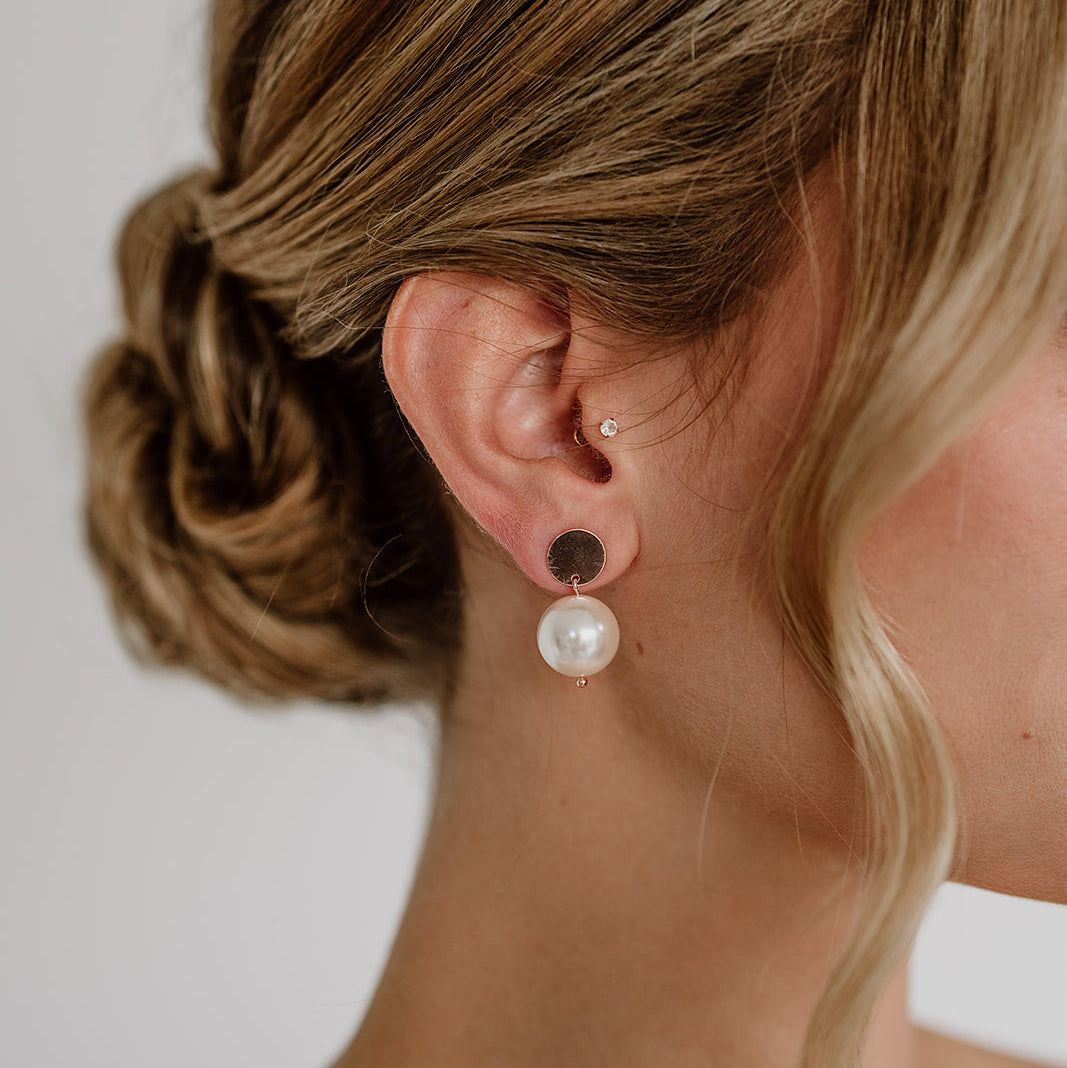 Zarte Brautohrringe aus 925er Sterlingsilber 18K rosévergoldet mit runder veganer Perle unter einem schlichten, runden Plättchen Ohrstecker, elegant getragen von einem Model am Ohr und harmonisch kombiniert mit einer Brautfrisur