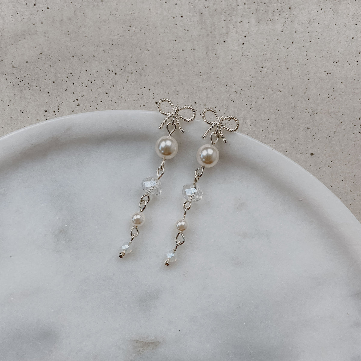 Brautohrringe Pauli mit Schleife aus 925 Sterlingsilber, darunter hängend Kristallperlen und veganer Perle, liegend auf einer Marmorplatte
