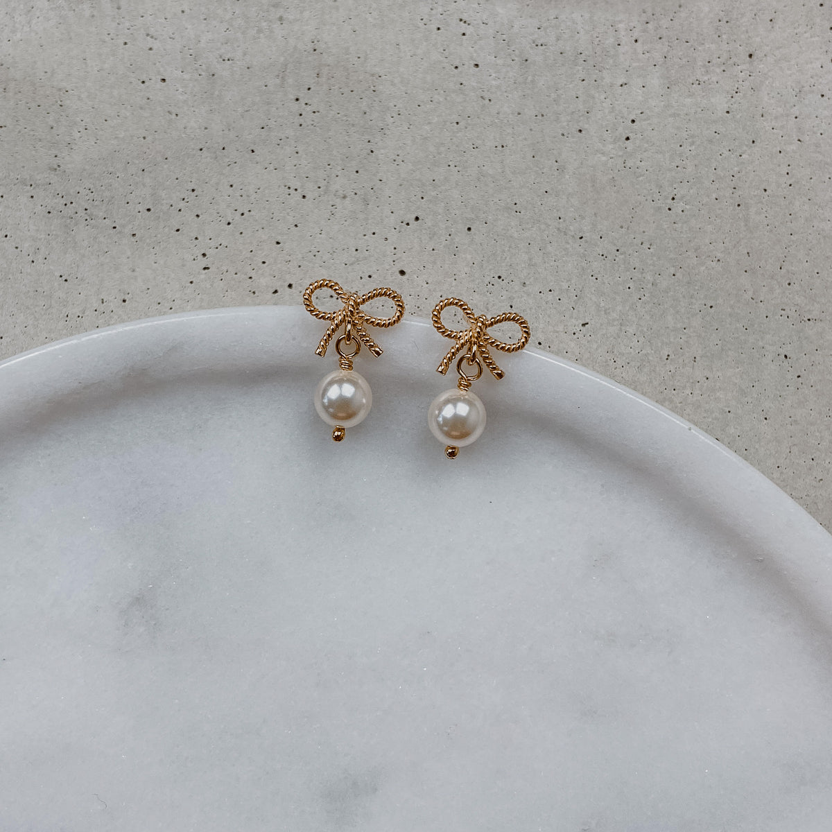 Braut Ohrringe Pinella, Schleifen Ohrstecker mit veganer Perle in 925 Sterlingsilber 18K rosévergoldet, liegend auf einer Marmorplatte