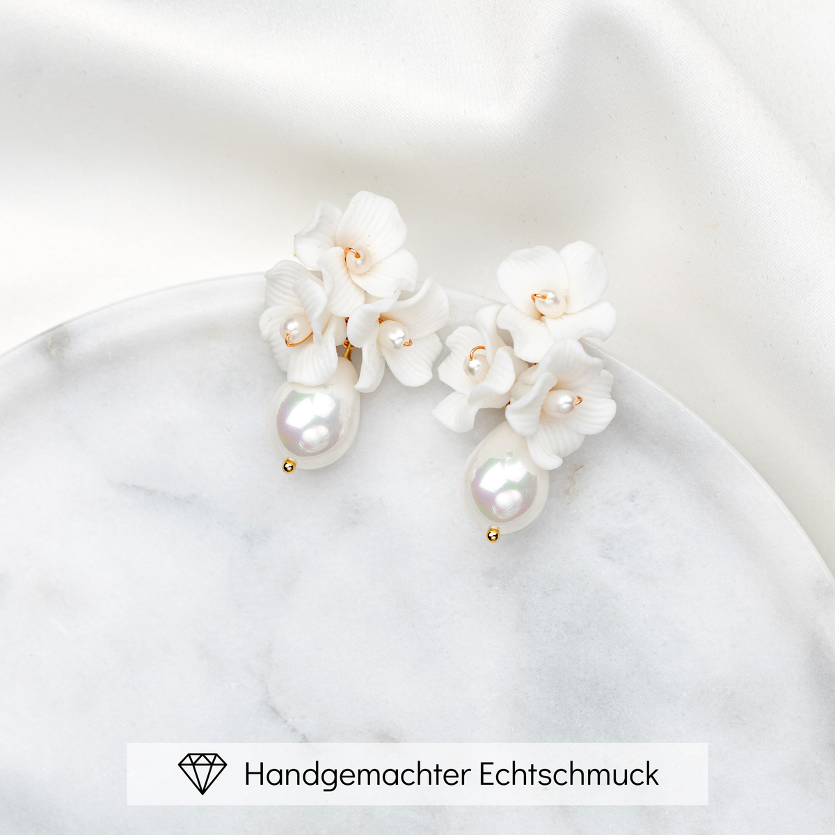 Elegante Brautohrringe aus 925er Sterlingsilber 18K vergoldet mit filigranen Porzellanblüten und kleinen Süßwasserperlen sowie darunter hängender tropfenförmiger Süßwasserperle, stilvoll arrangiert auf einer runden, hellen Marmorplatte.