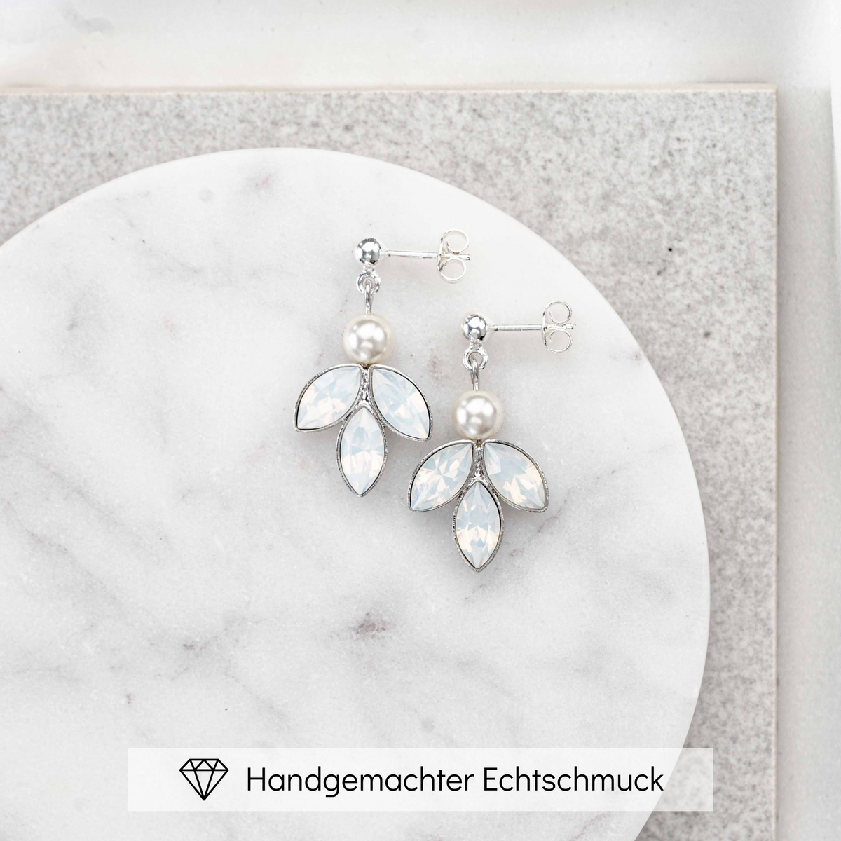 Zarte hängende Brautohrringe aus 925er Sterlingsilber mit rundem Kugelstecker, kleiner runder veganer Perle und darunter angeordneten blattförmigen, white opal Kristallen, ästhetisch arrangiert auf einer hellen Marmorplatte.