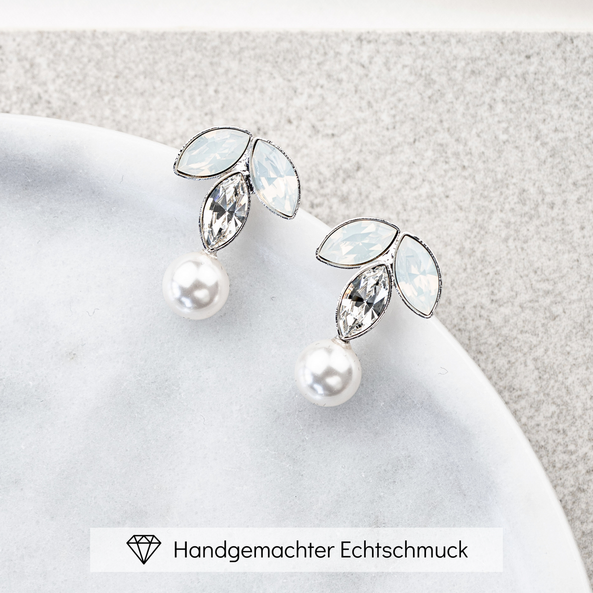 Brautohrringe Katy aus 925 Sterlingsilber mit white opal und crystal Kristallen in Blattform und veganer Perle, liegend auf einer Marmorplatte