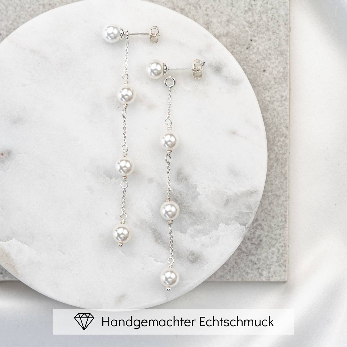 Zarte hängende Brautohrringe aus 925er Sterlingsilber mit rundem Perlenstecker und darunter in Reihe angeordneten, veganen, runden Perlen, verbunden mit einem feinen Kettchen, liegend auf einer hellen Marmorplatte. Der Perlenohrstecker ist abnehmbar und einzeln tragbar als schlichter Perlenstecker.