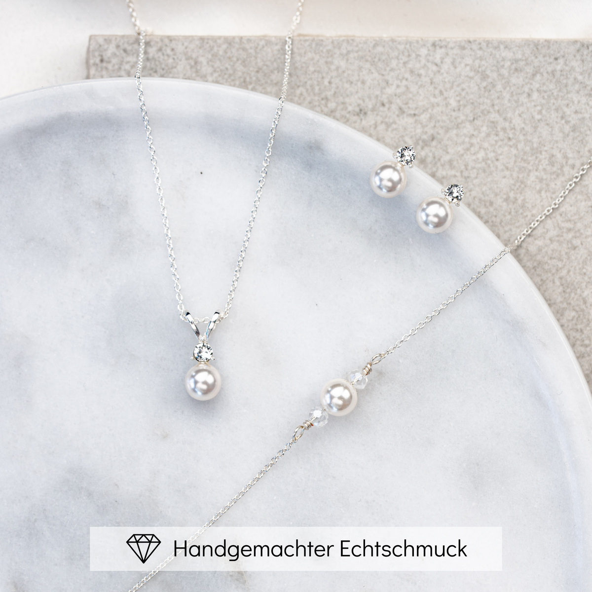 Hochwertiges Brautschmuck-Set aus 925er Sterlingsilber bestehend aus Brauthalskette, Ohrsteckern und Armband mit funkelndem Zirkonia Kristall und veganer Perle, ästhetisch arrangiert auf einer hellen Marmorplatte.