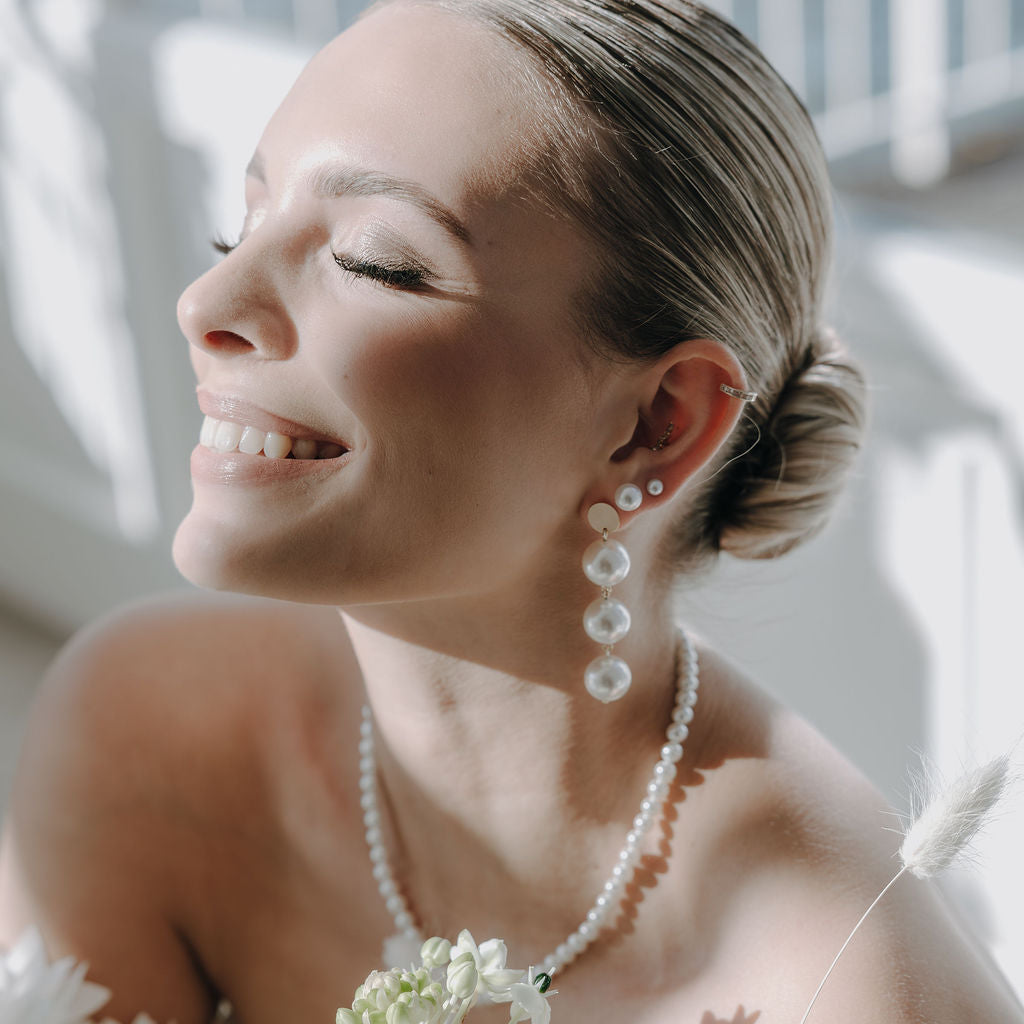 Elegante Brautohrringe aus 925er Sterlingsilber 18K vergoldet mit großen runden veganen Perlen untereinander hängend an einem schlichten, runden Plättchen Ohrstecker, stilvoll getragen von einem Model am Ohr und harmonisch kombiniert mit einer klassischen Perlenkette und Perlenohrstecker.