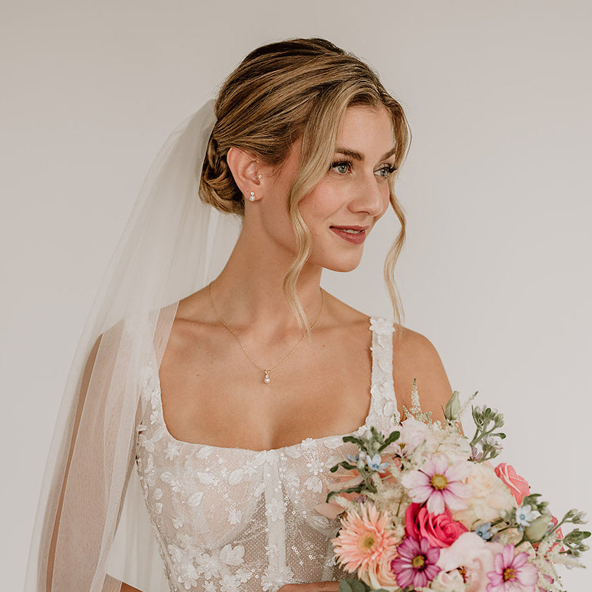 Stilvolle Brauthalskette aus 925er Sterlingsilber 18K vergoldet mit funkelndem Zirkonia Kristall und darunter hängender veganer Perle, detailreich am Hals eines Models mit Schleier getragen und harmonisch mit einer romantischen Brautfrisur kombiniert.