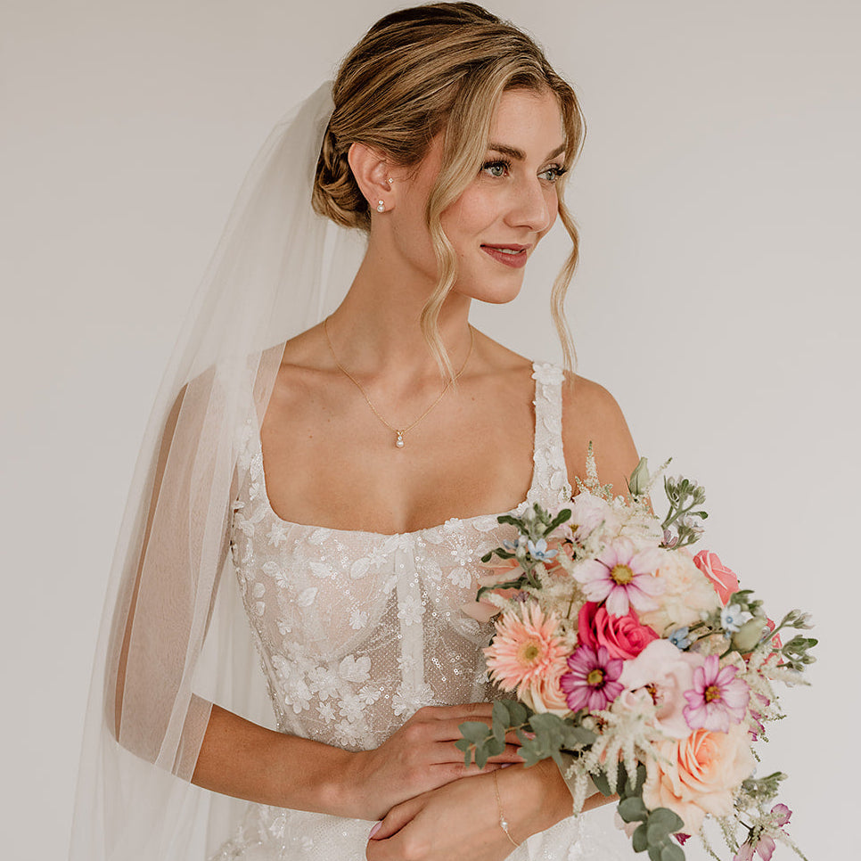Stilvolles Brautschmuck Set aus 925er Sterlingsilber 18K vergoldet mit funkelndem Zirkonia Kristall und darunter hängender veganer Perle, getragen von einem Model mit Schleier und harmonisch mit einer romantischen Brautfrisur kombiniert.