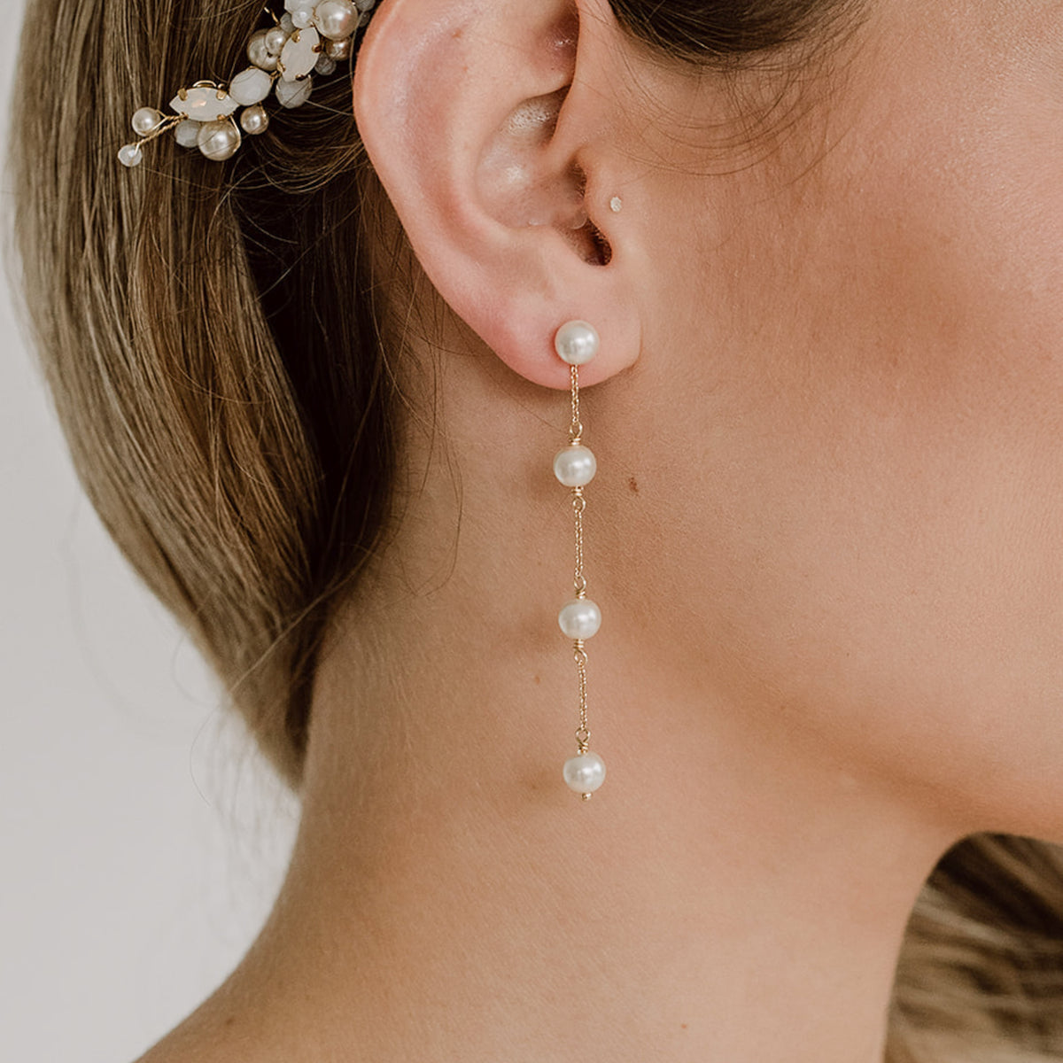 Zarte hängende Brautohrringe aus 925er Sterlingsilber 18K vergoldet mit rundem Perlenstecker und darunter in Reihe angeordneten, veganen, runden Perlen, verbunden mit einem feinen Kettchen, detailreich am Ohr eines Models mit eleganter Brautfrisur getragen.