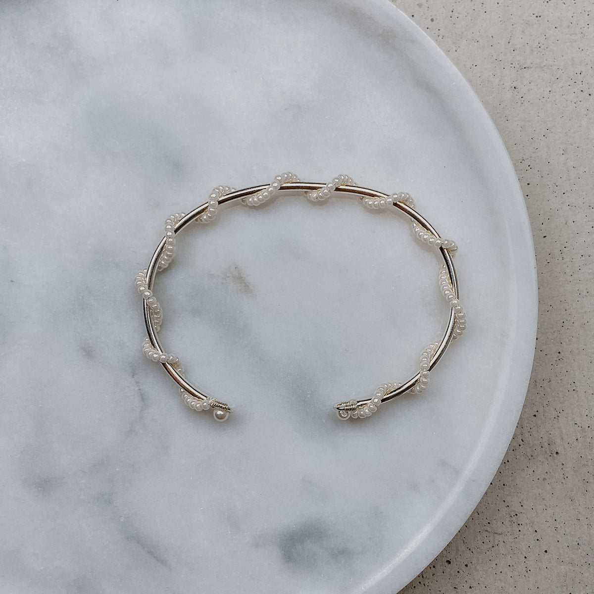 Braut Armreif Belina aus 925 Sterlingsilber mit kleinen Perlen, gewickelt um einen zarten Armreif, liegend auf Marmorplatte