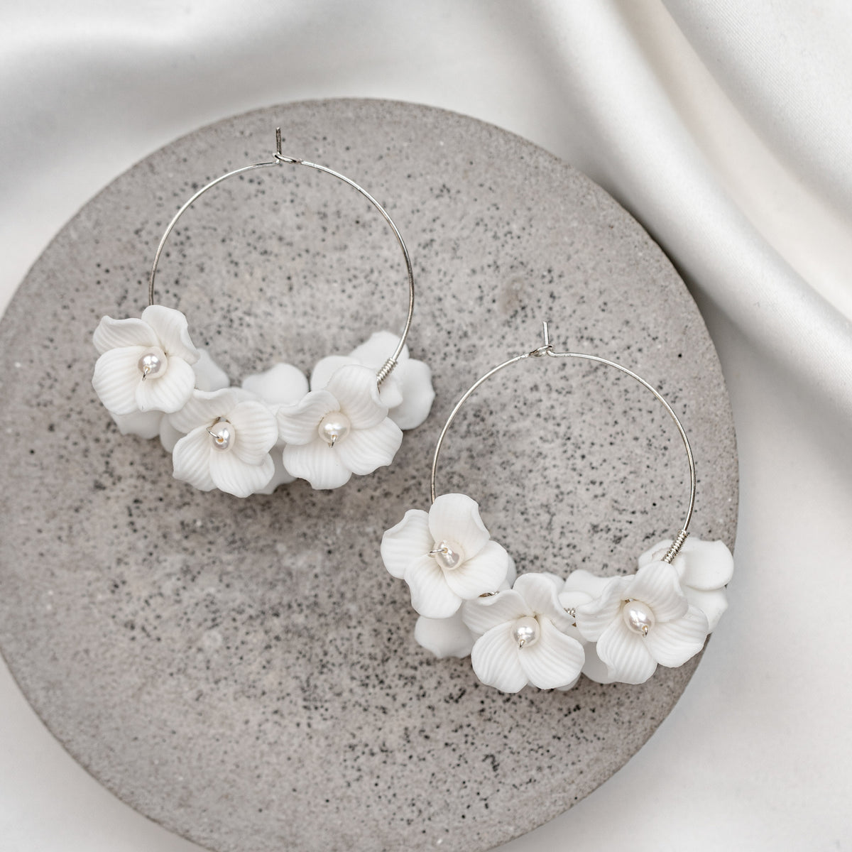 Elegante Brautohrringe aus 925er Sterlingsilber 18K vergoldet mit filigranen Porzellanblüten und kleinen Süßwasserperlen an einer feinen, runden Creole, stilvoll arrangiert auf einer grauen Betonoberfläche.