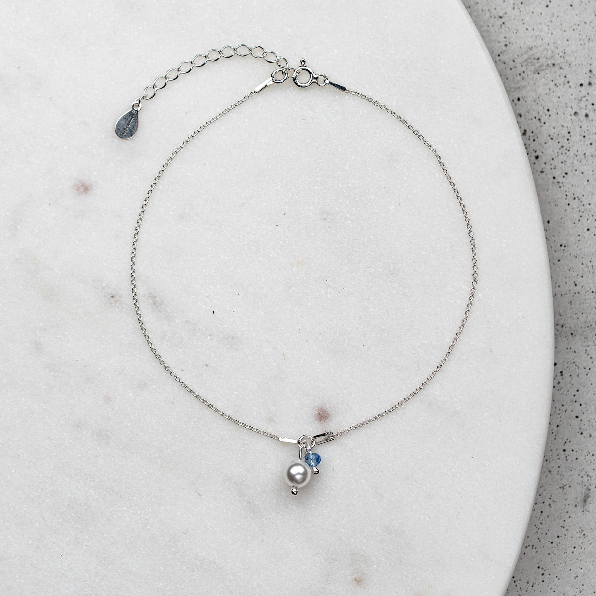 Braut Fußkettchen Saphira aus 925 Sterlingsilber  mit blauem Kristall und veganer Perle, liegend auf einer Marmorplatte