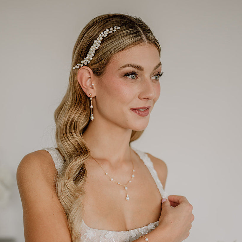 Zartes Brautschmuck-Set aus 925er Sterlingsilber 18K vergoldet bestehend aus hängenden Ohrringen mit unterschiedlich großen Süßwasserperlen sowie einer passenden Y-Halskette mit feinen Süßwasserperlen, elegant getragen von einem Model und kombiniert mit einem weißen, floral verzierten Brautkleid sowie dem passenden Haarschmuck.