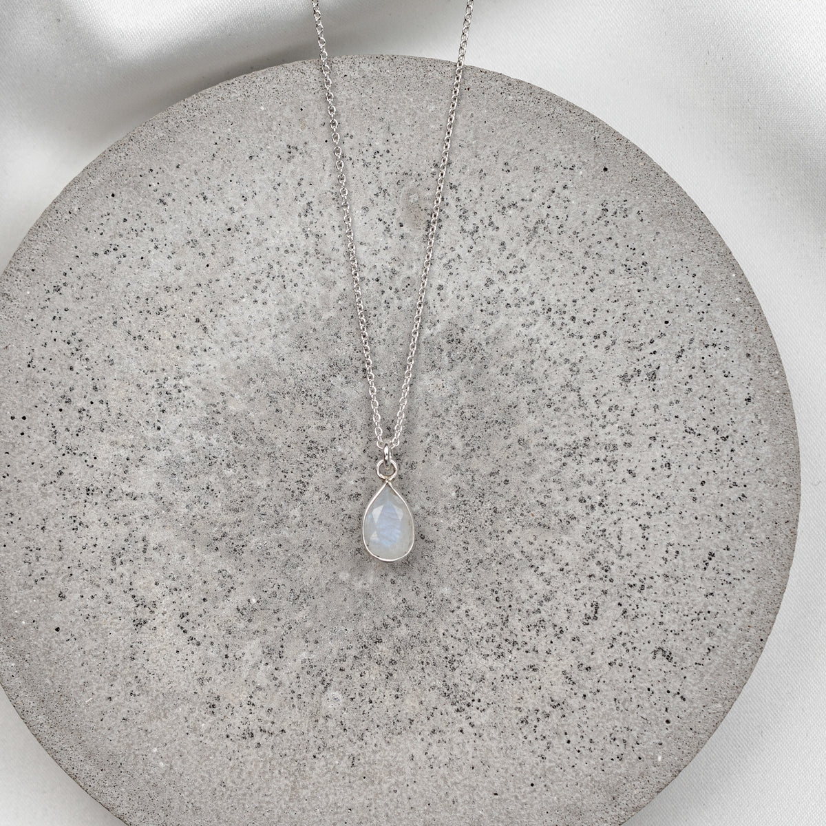 Braut Halskette aus 925er Sterlingsilber mit feiner Gliederkette und tropfenförmigem Mondstein Anhänger, präsentiert auf einer runden, hellgrauen Betonplatte