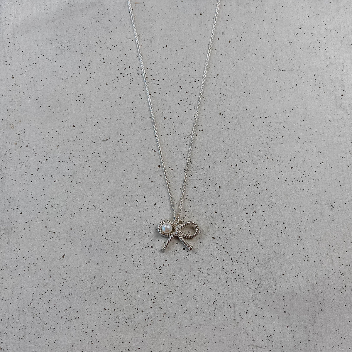 Braut Halskette Pinella aus 925 Sterlingsilber 18K vergoldet mit Schleife und veganer Perle, liegend auf Betonplatte