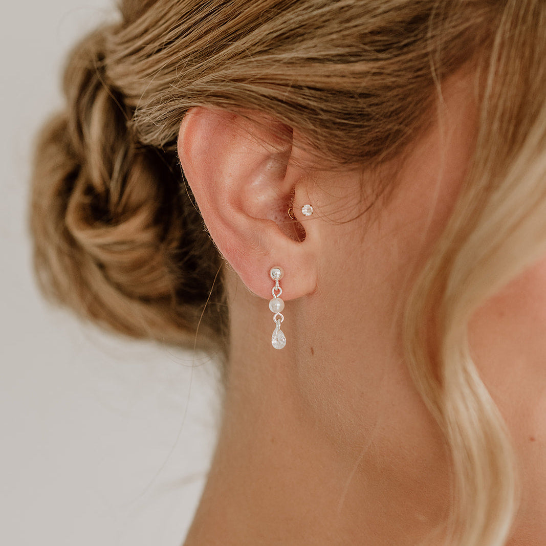 Zarte Brautohrringe aus 925er Sterlingsilber mit hängendem tropfenförmigem Zirkonia Kristall und kleiner runder Perle, elegant getragen von einem Model am Ohr und harmonisch kombiniert mit einer Hochsteckbrautfrisur.