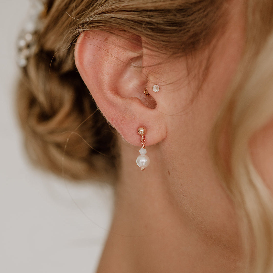 Feine Brautohrringe aus 925er Sterlingsilber 18K rosévergoldet. Kugelohrstecker mit kleiner Kristallperle und darunter hängender runder Perle, elegant getragen von einem Model am Ohr und harmonisch kombiniert mit einer klassischen Brautfrisur.