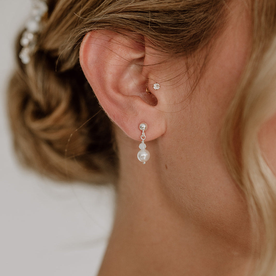 Feine Brautohrringe aus 925er Sterlingsilber. Kugelohrstecker mit kleiner Kristallperle und darunter hängender runder Perle, elegant getragen von einem Model am Ohr und harmonisch kombiniert mit einer klassischen Brautfrisur.