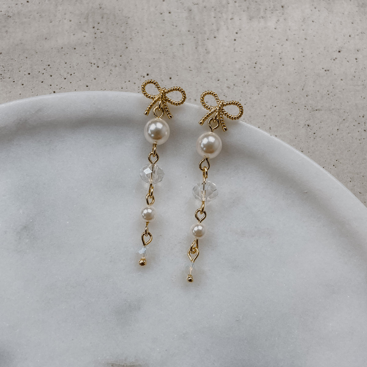 Brautohrringe Pauli mit Schleife aus 925 Sterlingsilber 18K vergoldet, darunter hängend Kristallperlen und veganer Perle, liegend auf einer Marmorplatte