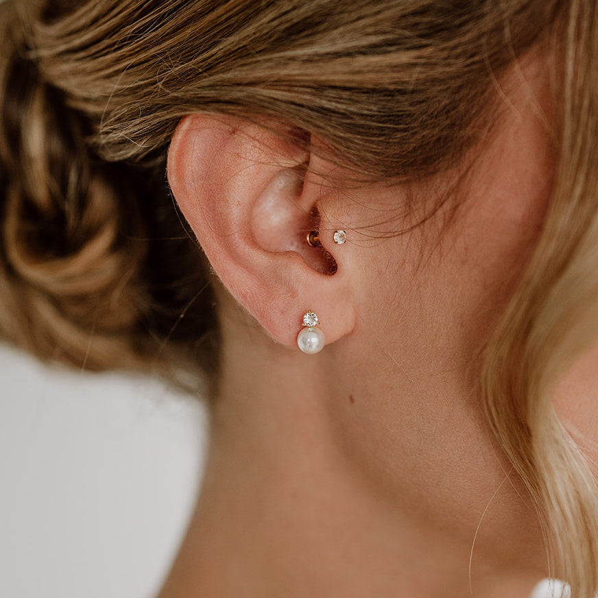 Feine Brautohrstecker aus 925er Sterlingsilber 18K vergoldet mit funkelndem Zirkonia Kristall und darunter sitzender runder veganer Perle, stilvoll am Ohr eines Models getragen und harmonisch mit einer klassischen Brautfrisur kombiniert.