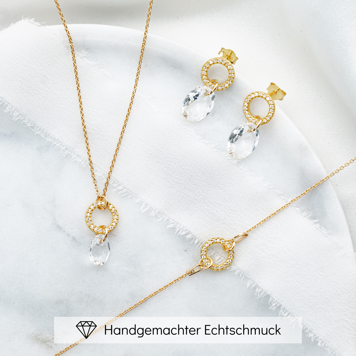 3-teiliges Brautschmuckset Mimi aus 925 Sterlingsilber 18K vergoldet – Ohrringe und Halskette mit rundem Element und Zirkonia Kristallen, darunter hängend ein facettierter Kristall, Armband mit rundem Element und Zirkonia Kristallen, liegend auf Marmorplatte