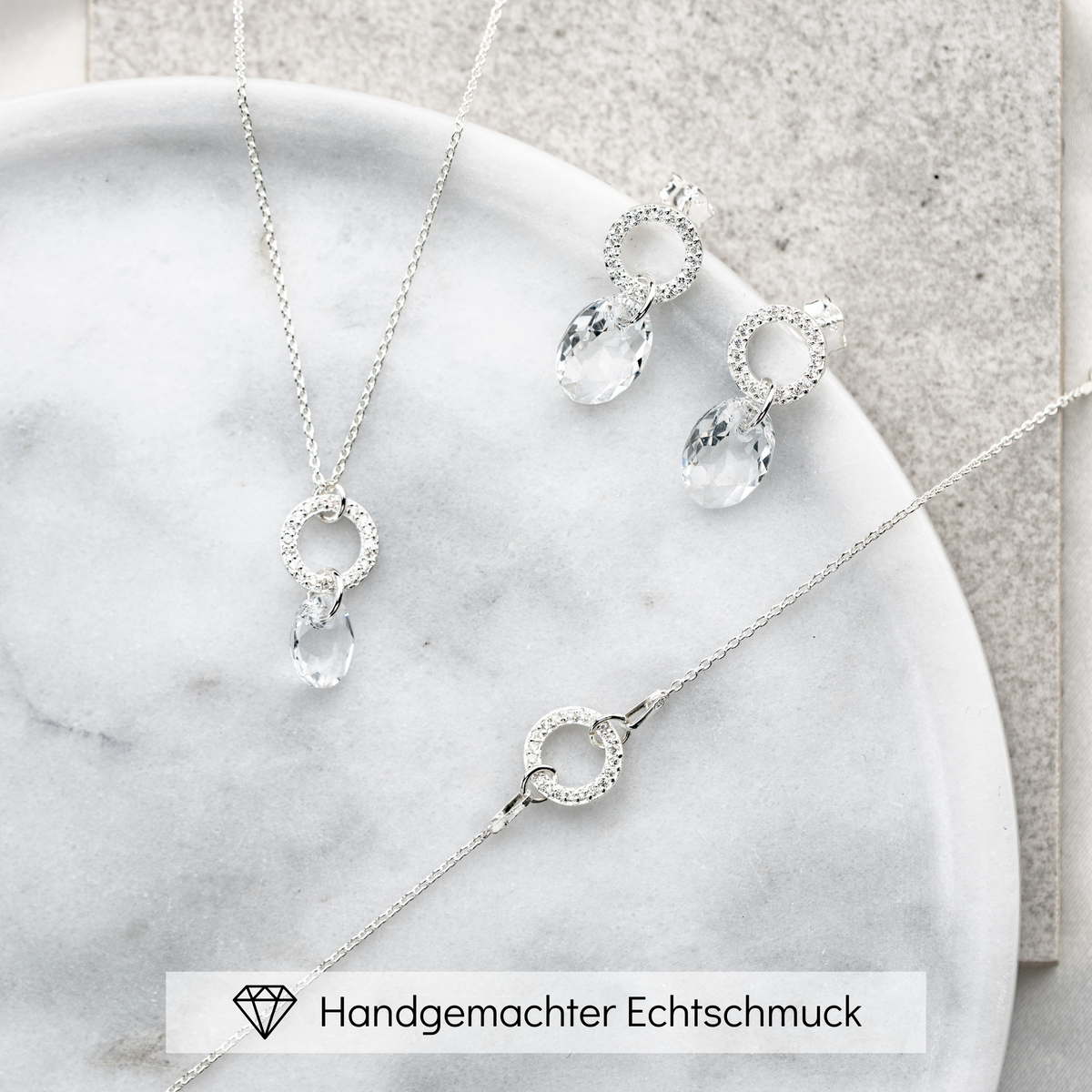 3-teiliges Brautschmuckset Mimi aus 925 Sterlingsilber – Ohrringe und Halskette mit rundem Element und Zirkonia Kristallen, darunter hängend ein facettierter Kristall, Armband mit rundem Element und Zirkonia Kristallen, liegend auf Marmorplatte