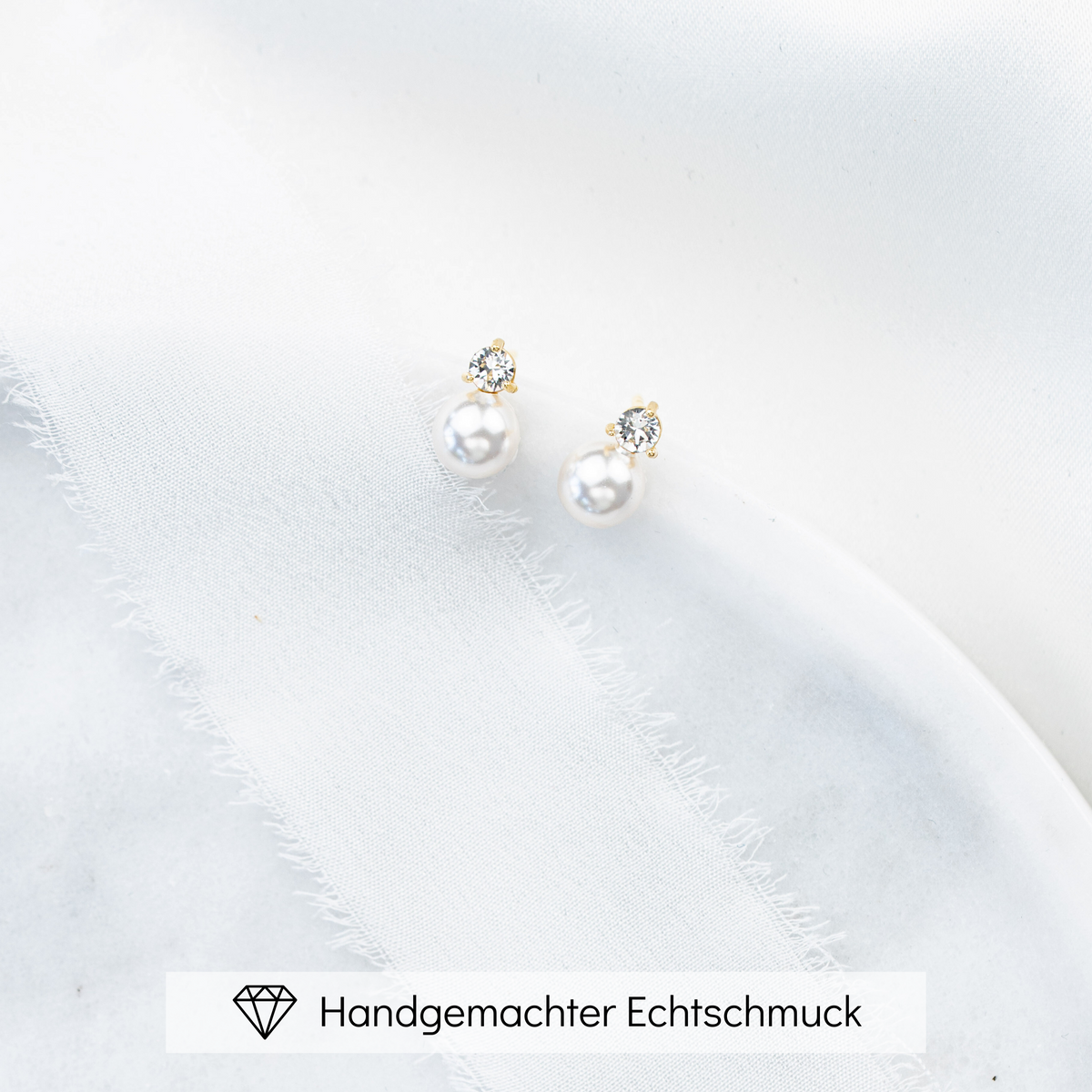 Feine Brautohrstecker aus 925er Sterlingsilber 18K vergoldet mit funkelndem Zirkonia Kristall und darunter sitzender runder veganer Perle, ästhetisch arrangiert auf einer hellen Marmorplatte.