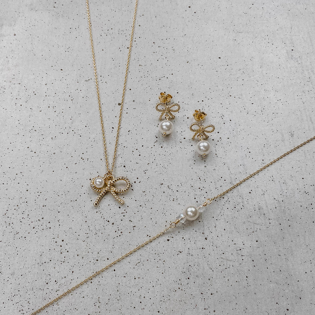 Braut Ohrringe Pinella, Schleifen Ohrstecker mit veganer Perle in 925 Sterlingsilber 18K vergoldet mit passender Braut Halskette Pinella und Braut Armband Ari, liegend auf einer Betonplatte