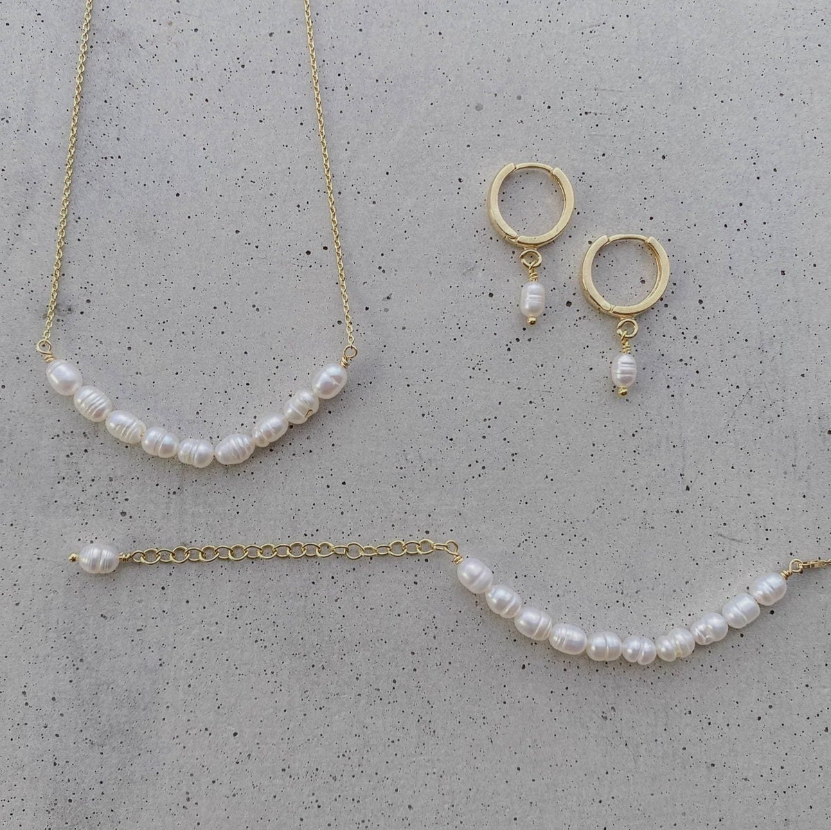 Braut Armband Linn aus 925 Sterlingsilber 18K vergoldet mit echten Süßwasserperlen mit passenden Braut Creolen Amelie und Braut Halskette Linn, liegend auf einer Betonplatte