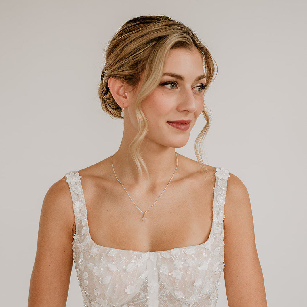 Zarte Brauthalskette Juna aus 925er Sterlingsilber mit kleinem tropfenförmigem Mondstein Anhänger, kombiniert mit passenden Mondstein Ohrringen Juna, elegant getragen von einem Model und abgestimmt auf ein weißes, floral verziertes Brautkleid.