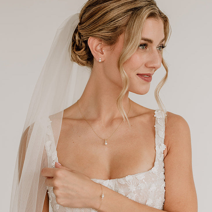 Stilvolles Brautschmuck-Set aus 925er Sterlingsilber 18K vergoldet bestehend aus Brauthalskette, Ohrsteckern und Armband mit funkelnden Zirkonia Kristallen und veganen Perlen, harmonisch an einem Model mit Schleier getragen und elegant mit einer klassischen Brautfrisur kombiniert.